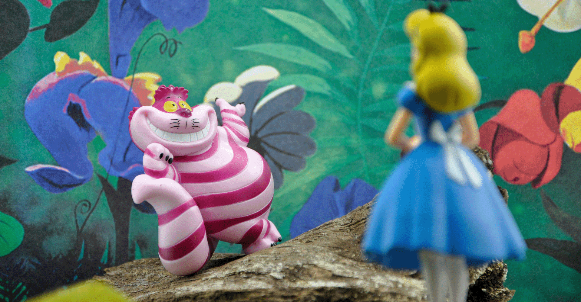 Alice-Figur steht vor dem grinsenden Kater aus Alice im Wunderland, beide umgeben von einer farbenfrohen Fantasielandschaft.